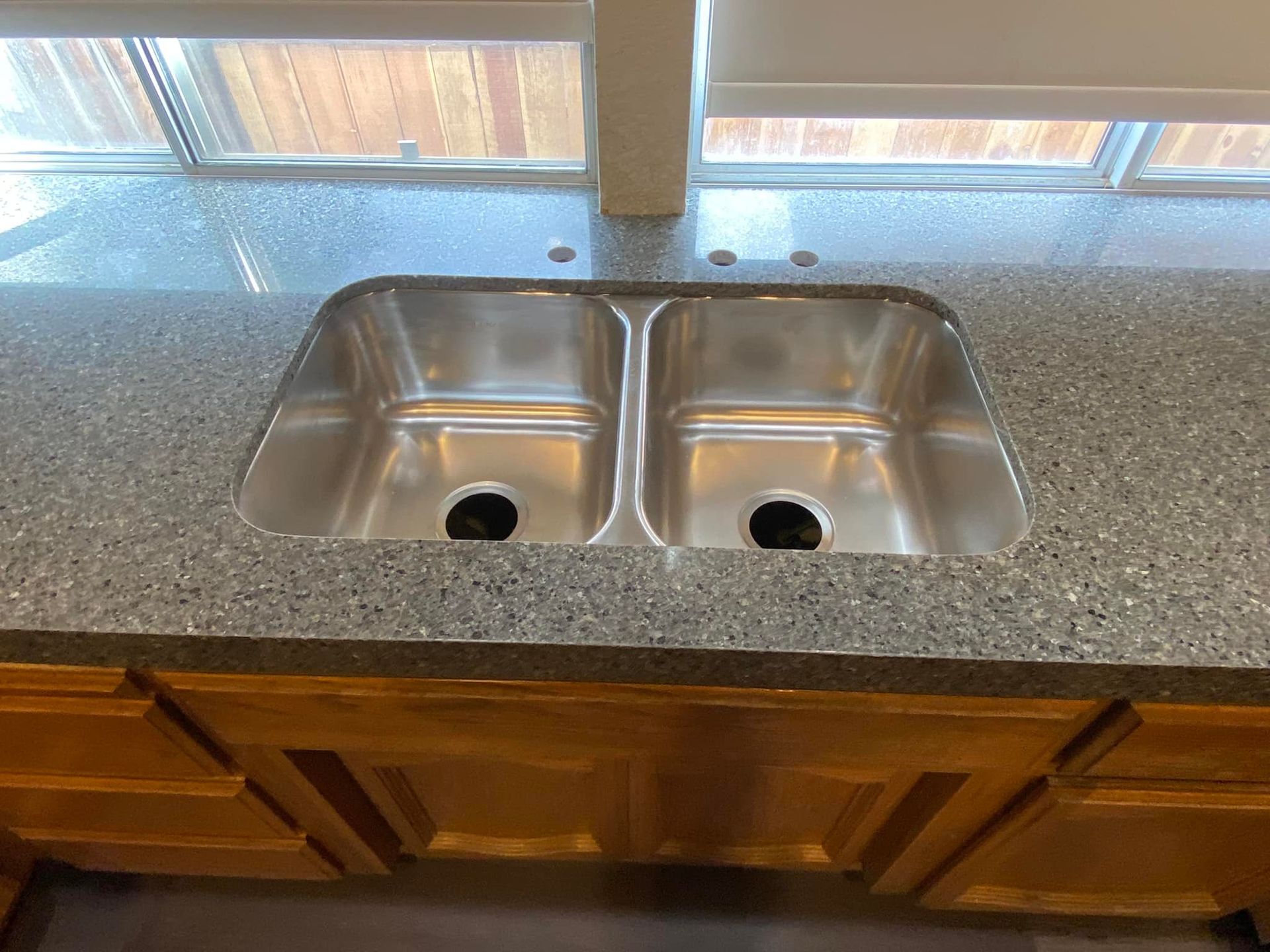 Stainless steel double sink in a speckled gray countertop, with wood cabinets below and a window above.