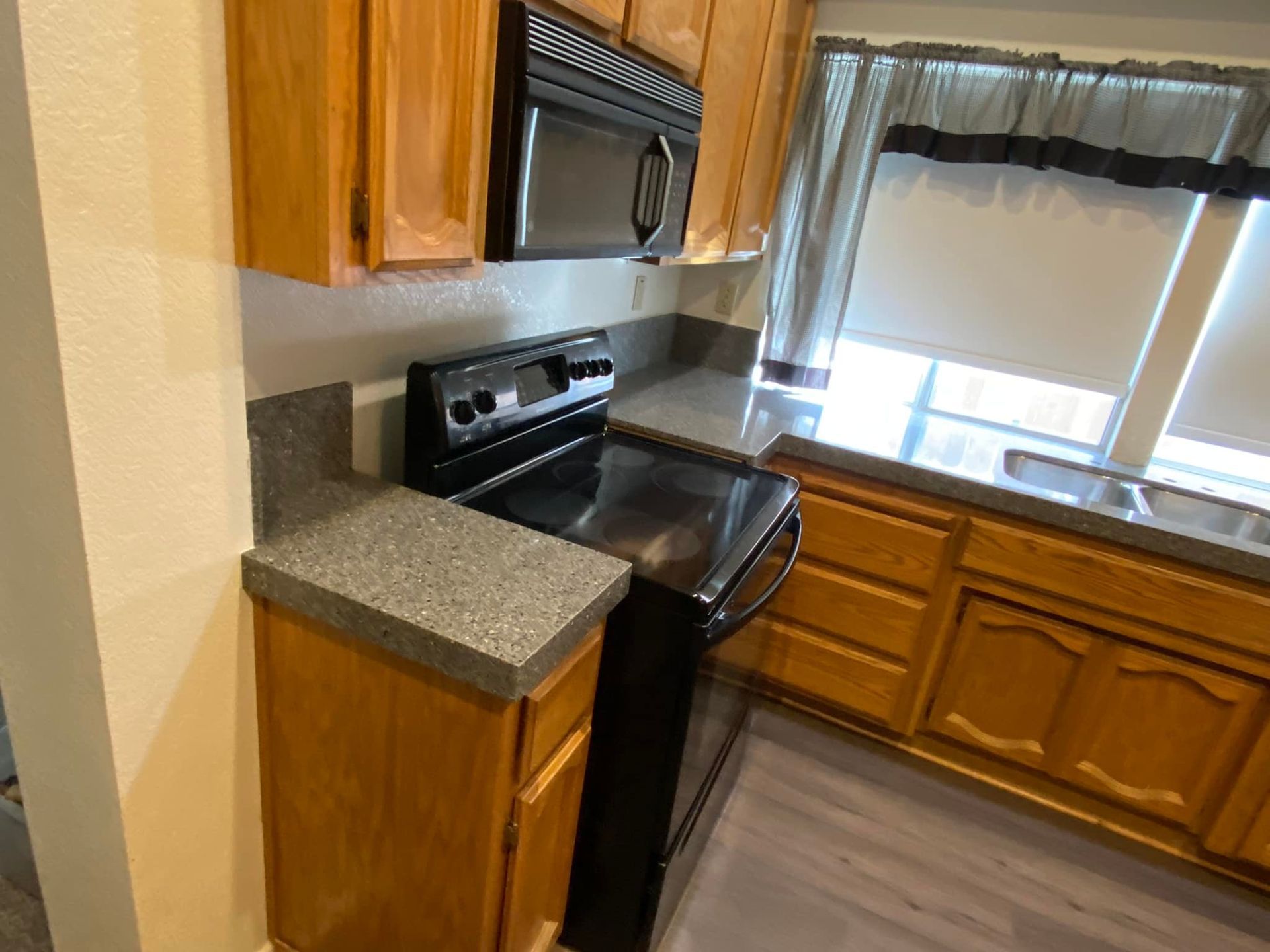 Kitchen with wooden cabinets, black microwave and stove, gray countertops, and a window.