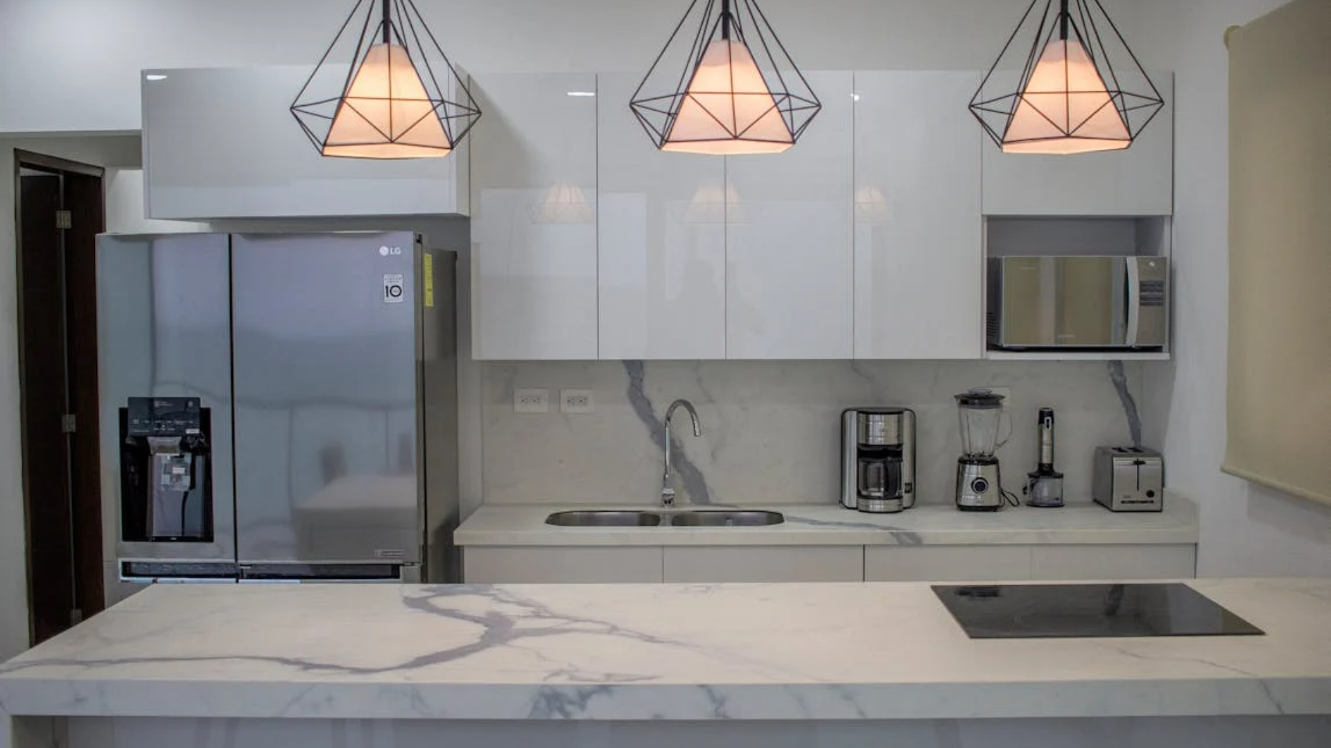 Modern white kitchen with stainless steel appliances, marble countertops, and geometric pendant lights.