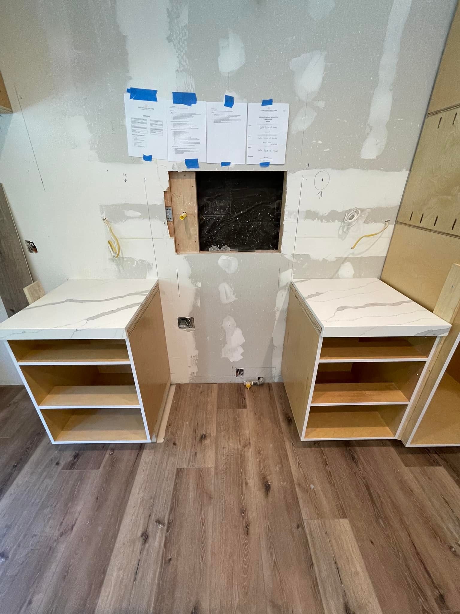Two open kitchen cabinets with countertops flank a hole in a wall; construction site.