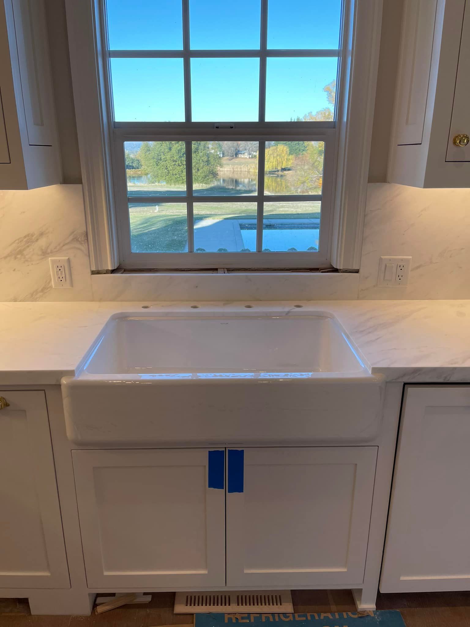 White farmhouse sink under a window in a kitchen. White cabinets and countertops. Blue tape on doors.