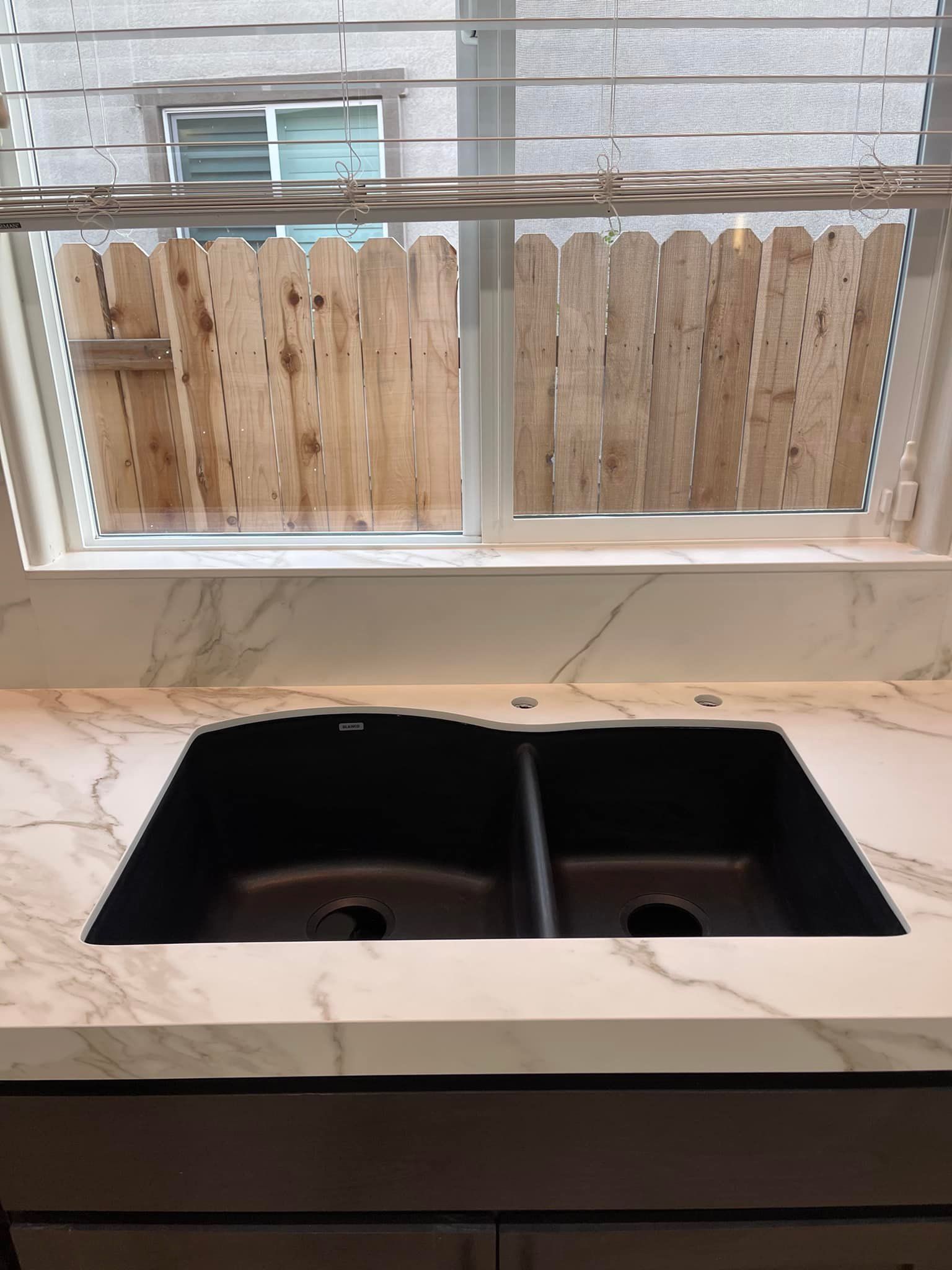 A black double sink in a marble-patterned countertop, under a window with a wooden fence view.