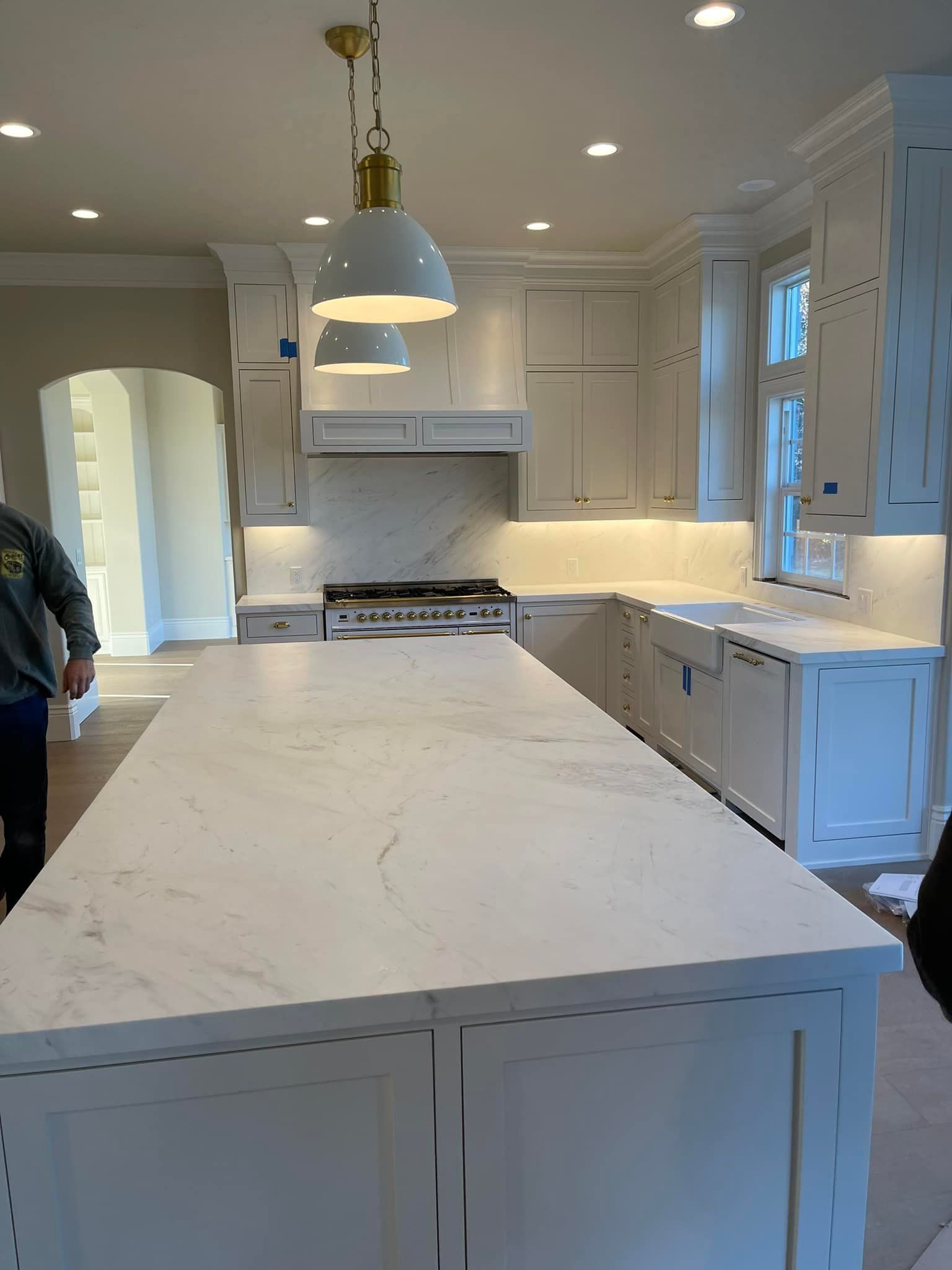 White kitchen with island, cabinets, marble backsplash, pendant lights, and person.