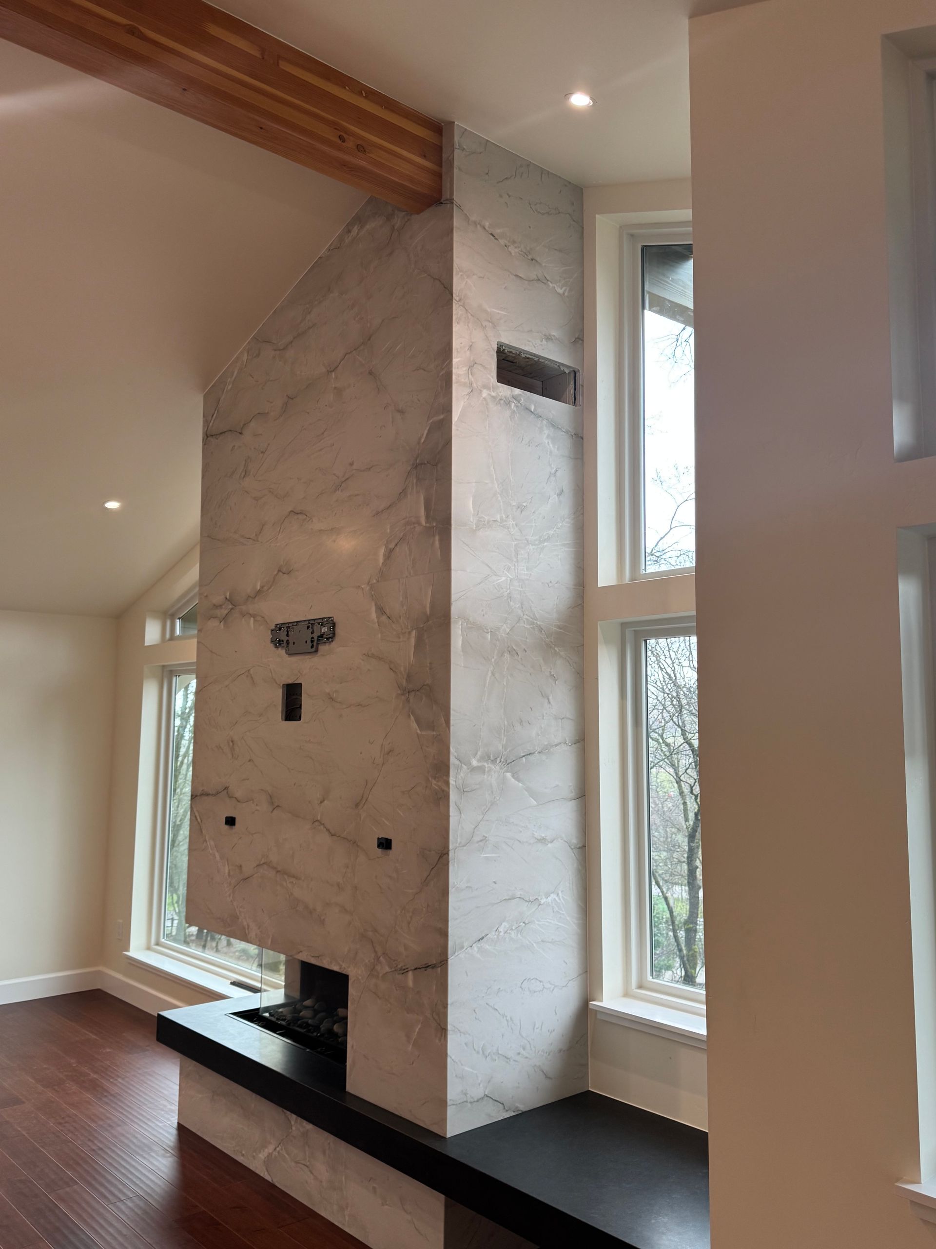 A modern fireplace with a marble facade, flanked by windows, dark hardwood floors, and a wooden beam.