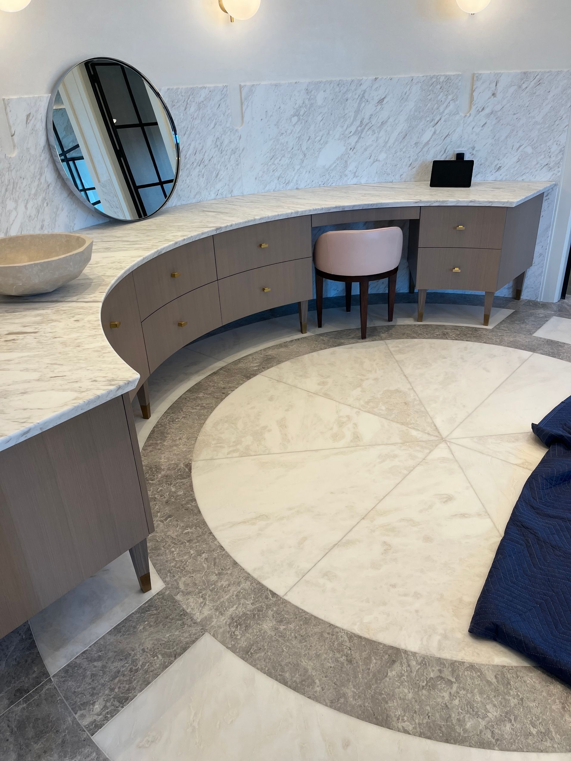 Elegant vanity with round mirror and pink stool in a luxury bathroom with marble floors.