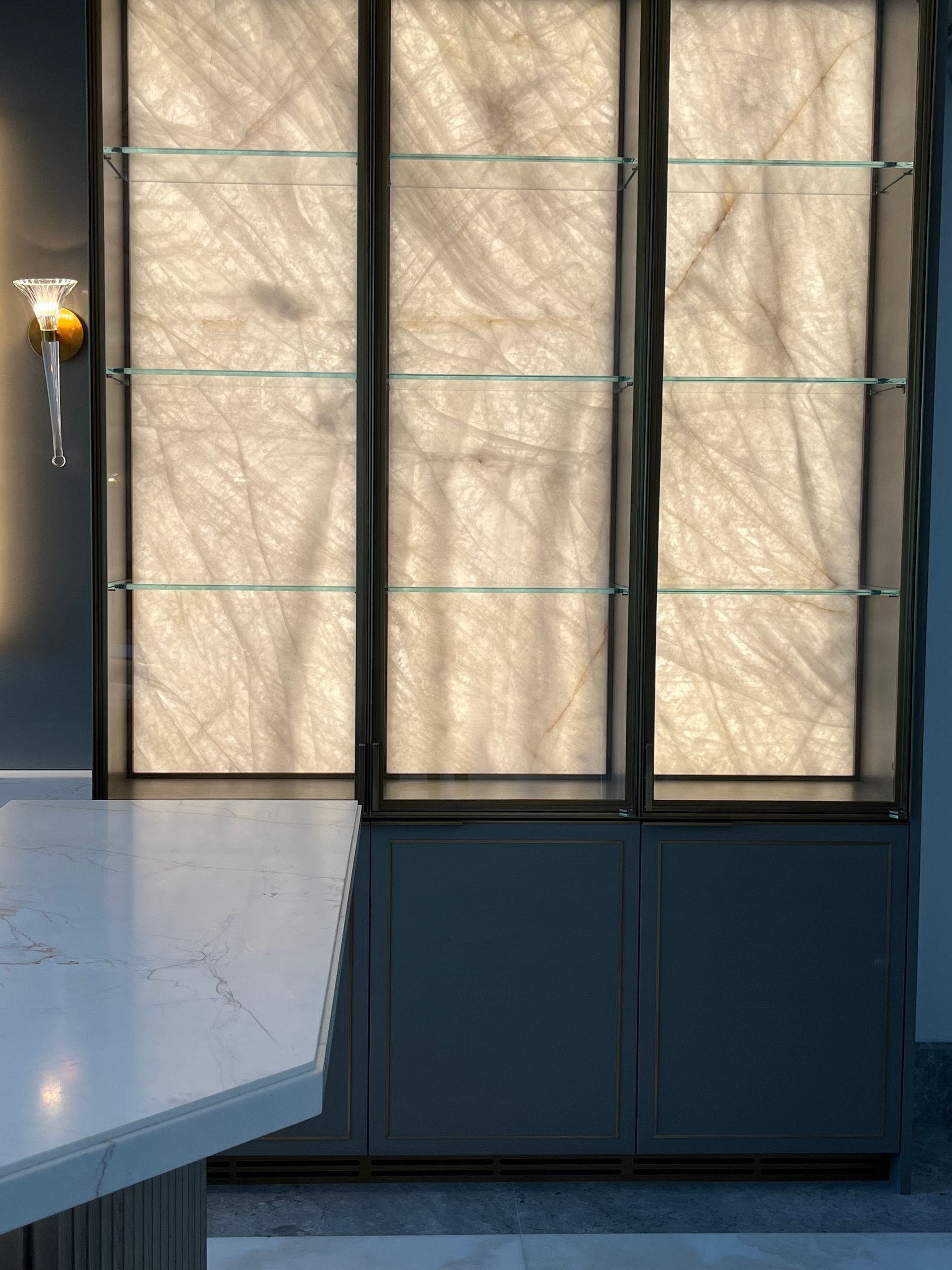 Lit display cabinet with three glass shelves against a textured backdrop, with a countertop and a lamp.