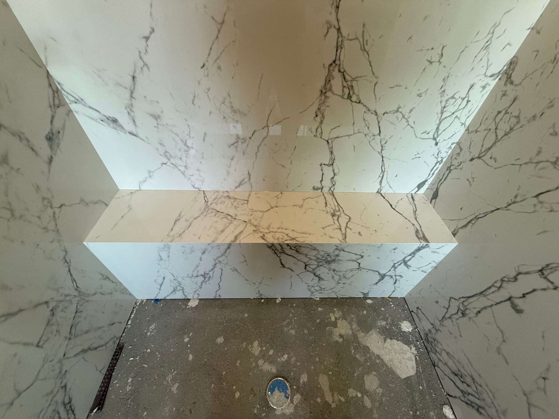 Shower interior with marble-patterned walls and a built-in shelf; unfinished concrete floor.