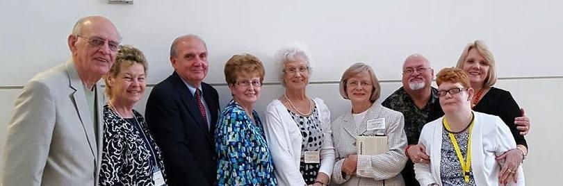 Group of eight people posing indoors, smiling in formal and patterned clothing.