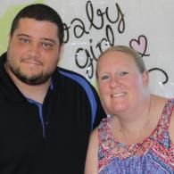 Two people smiling in front of a wall with “baby girl” and a heart decoration