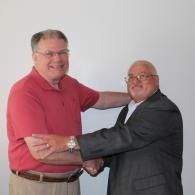 Two men shaking hands indoors, one in a red shirt and one in a dark suit, smiling.