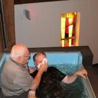 Person assists another in a blue baptismal pool inside a church, with a stained-glass window behind them.