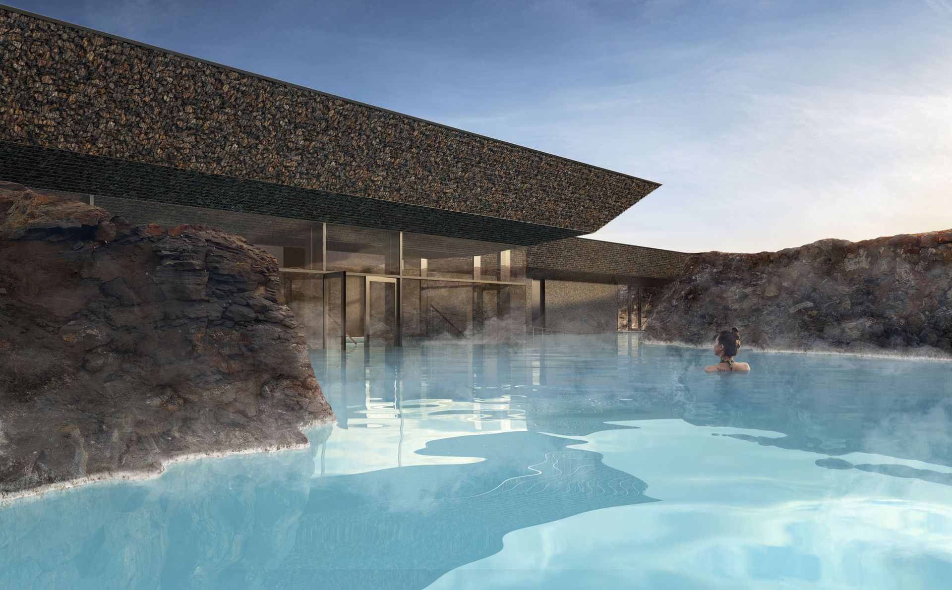 A person in a turquoise thermal pool near a modern building with stone exterior in a natural landscape.