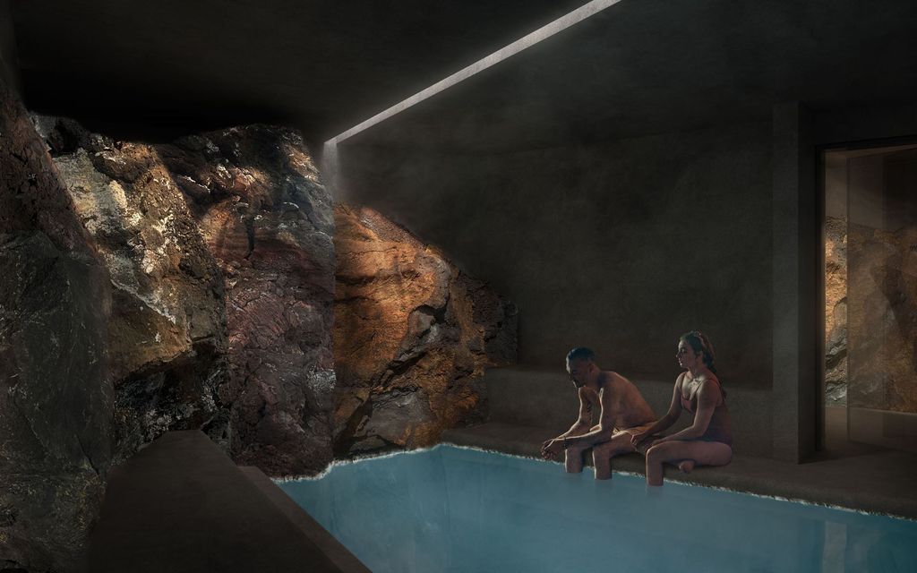 Two people sit by an indoor pool in a dim cave-like room with rocky walls and a skylight.