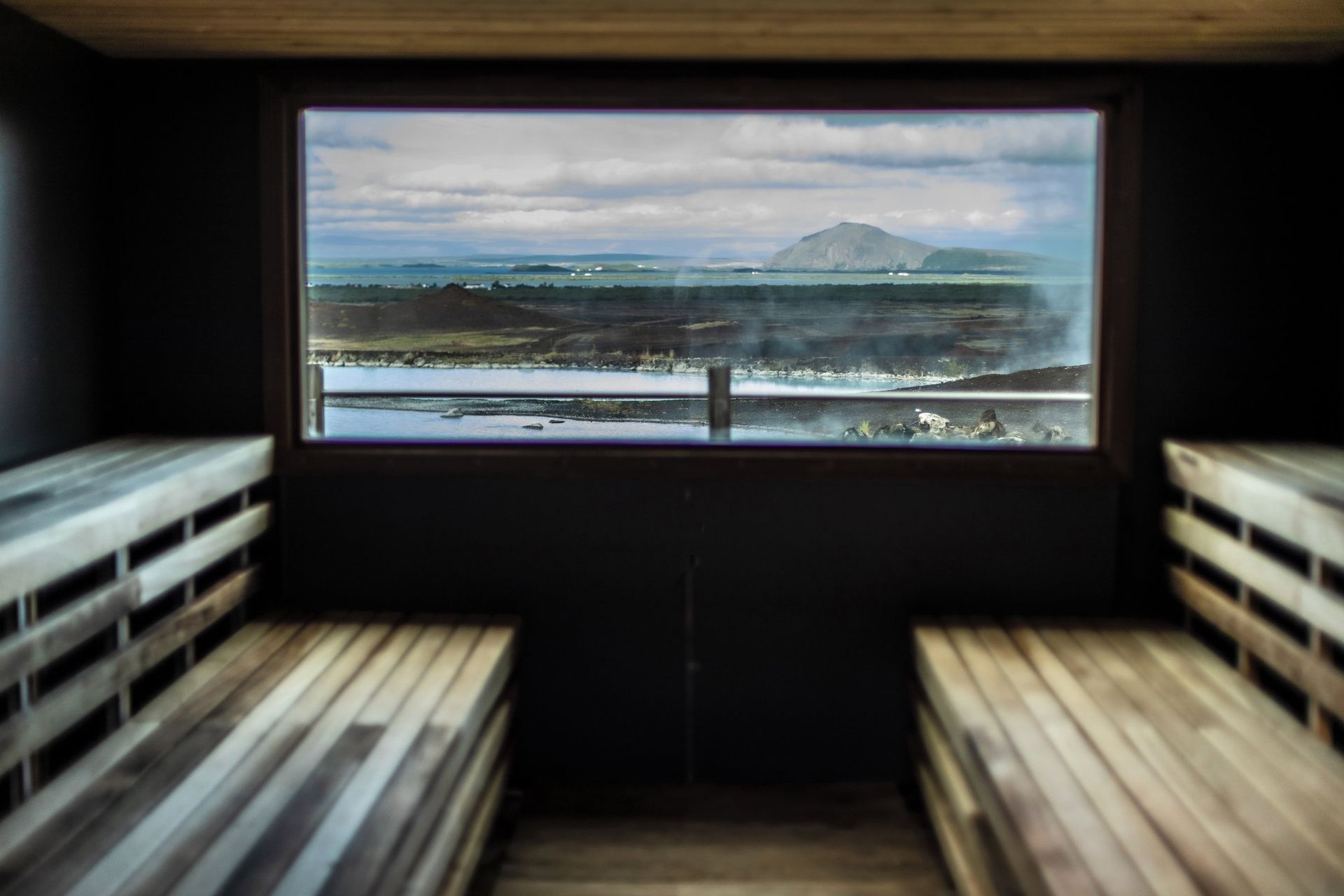 Wooden sauna interior with a window overlooking a vast landscape under a cloudy sky.
