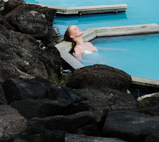 Woman with red hair relaxes in blue geothermal spa water, near dark rocks.