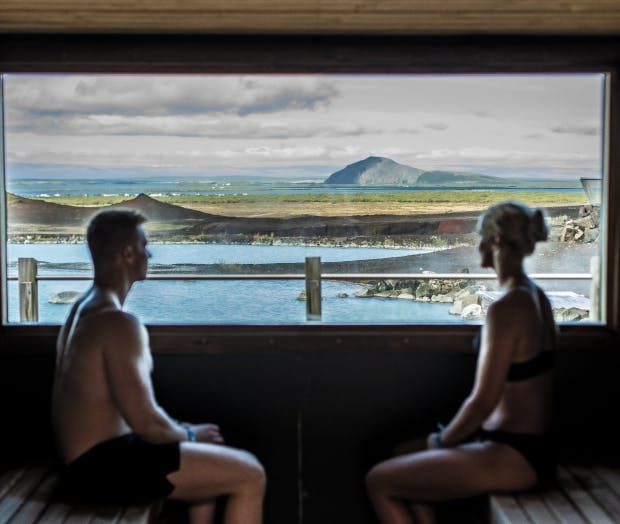 Par í gufubaði og horfir yfir landslag með vatni og fjöllum.