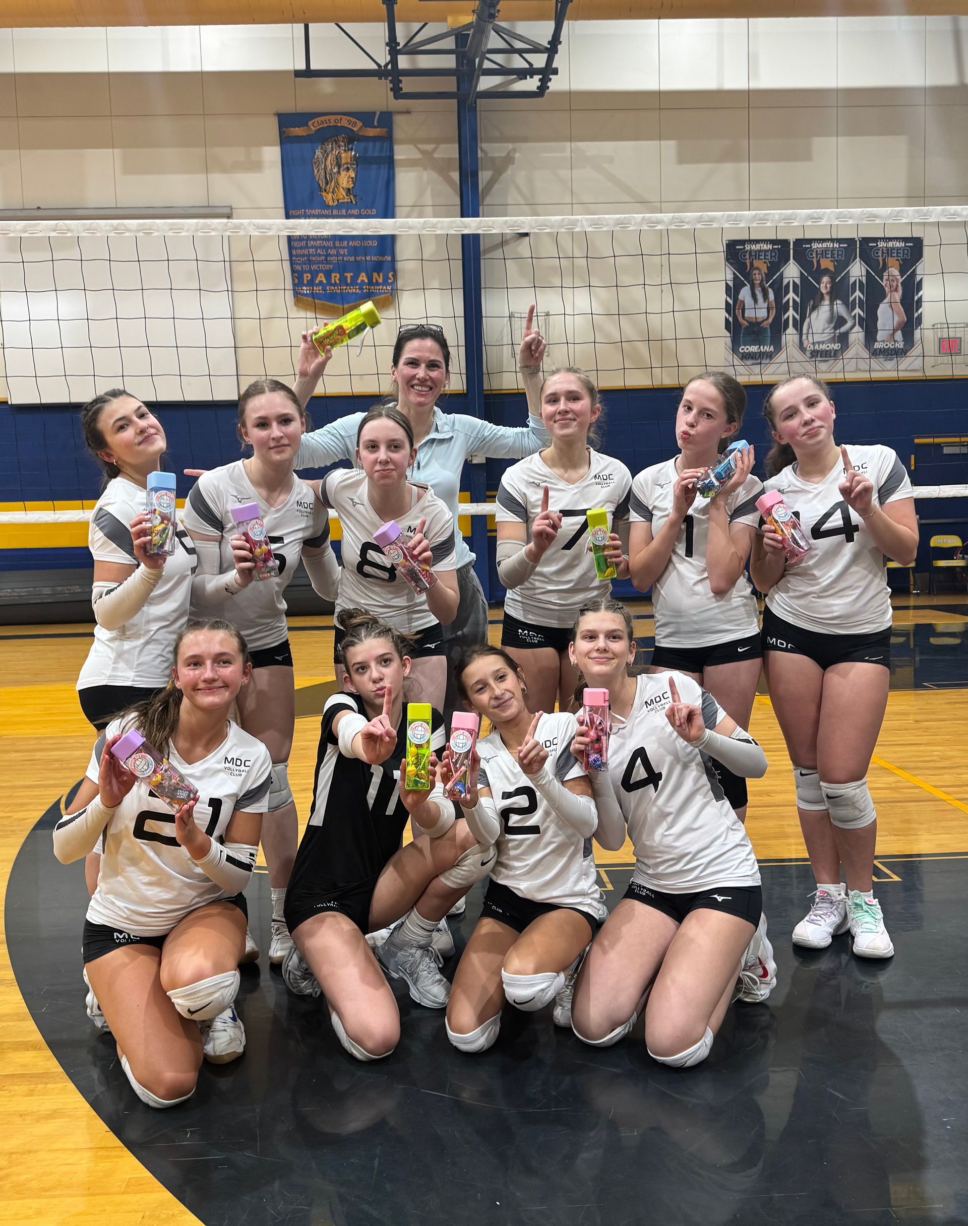 Volleyball team holding snacks, posing on court, wearing white and black uniforms, smiles.
