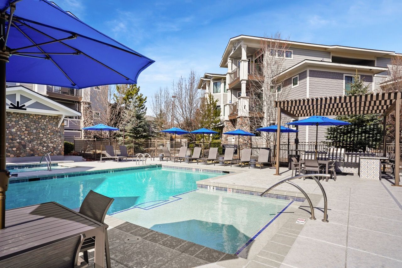 Outdoor community pool with lounge chairs and blue umbrellas near apartment buildings.