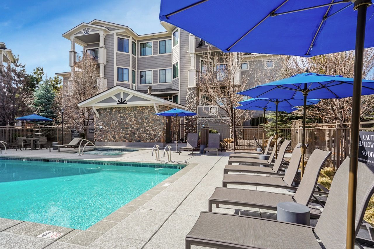Outdoor pool area at an apartment complex with lounge chairs, blue umbrellas, and a stone poolhouse.