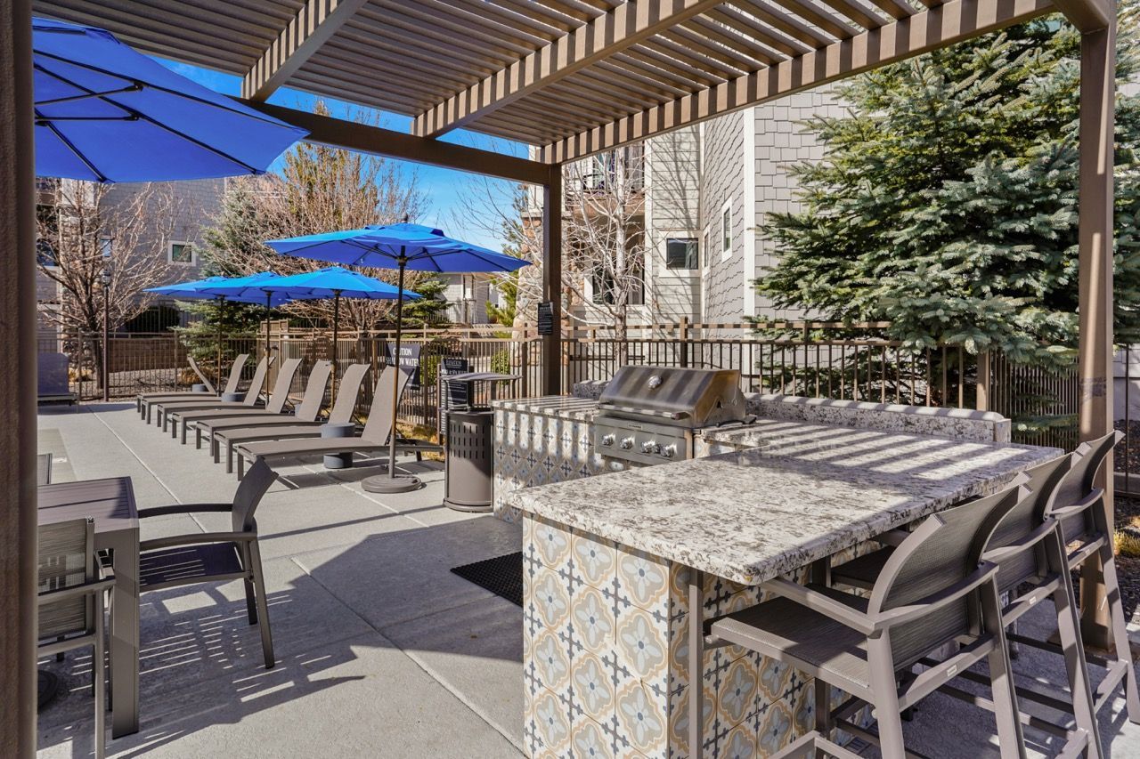 Outdoor apartment amenity area with a pergola, blue umbrellas, lounge chairs, and a grilling station.