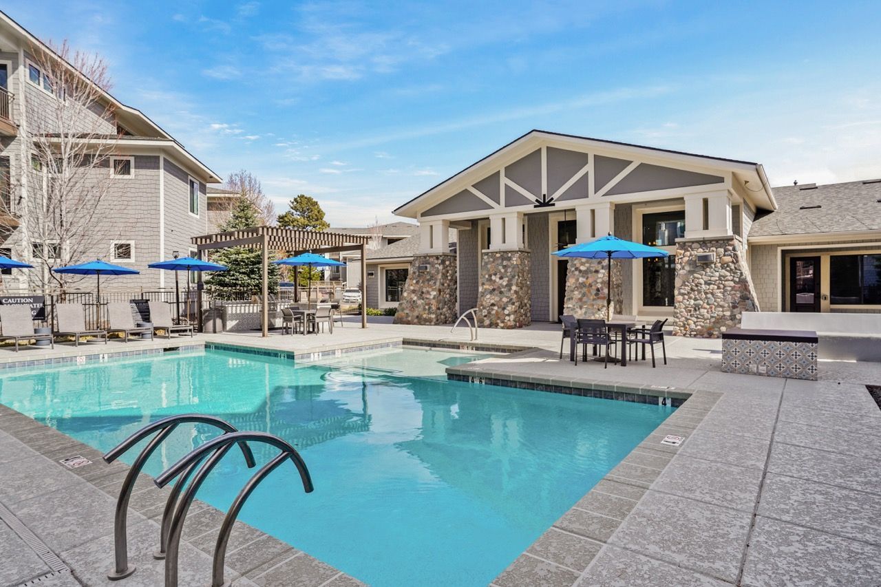 Outdoor apartment community pool with lounge chairs, blue umbrellas, and a clubhouse.