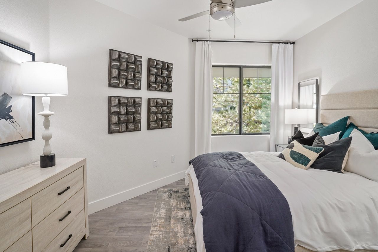 Bedroom in a modern apartment with a bed, dresser, and a window with white curtains.