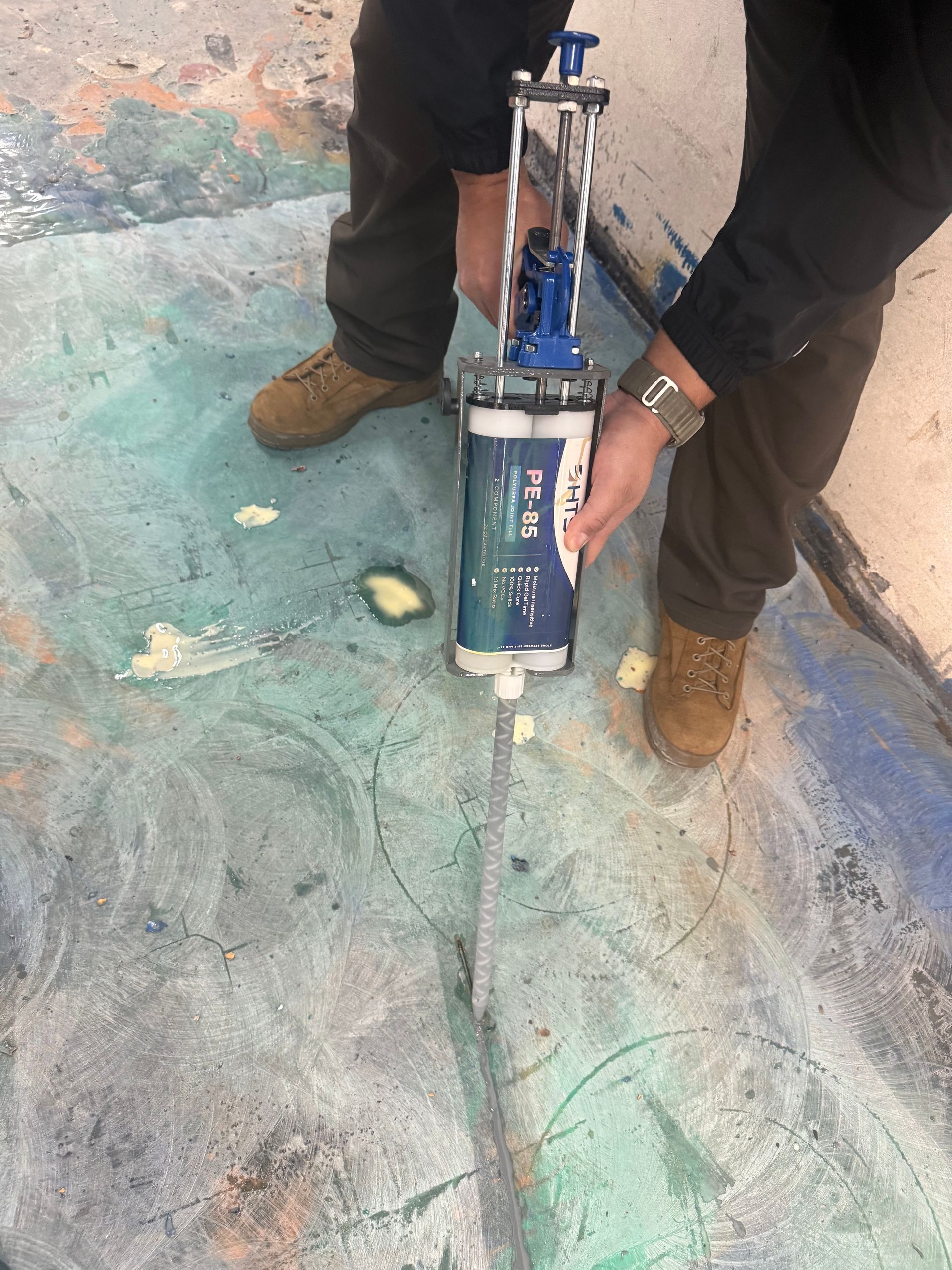 Person in blue jacket and jeans spreading gray sealant on a floor with a tool.