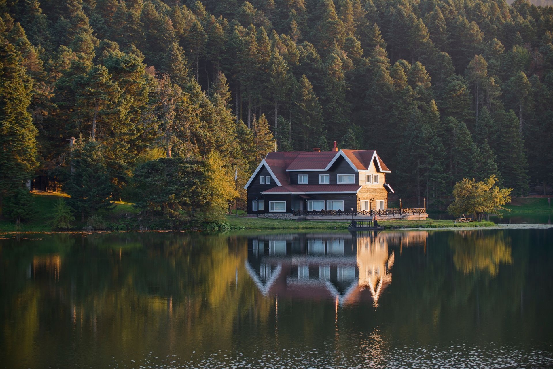 A house sits lakeside, reflecting in the water, surrounded by a dense forest.