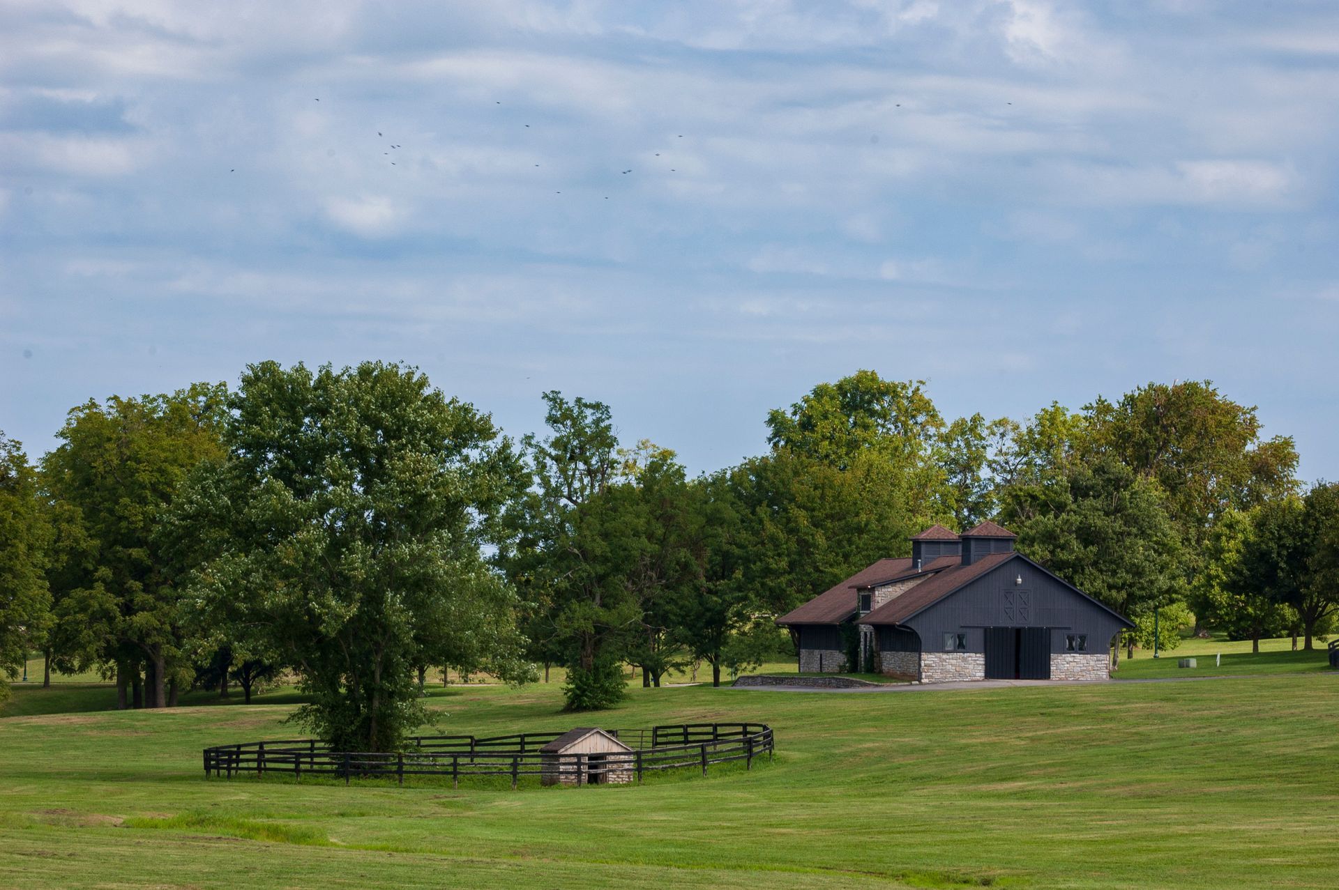 Barn in a grassy field, surrounded by trees under a cloudy sky.