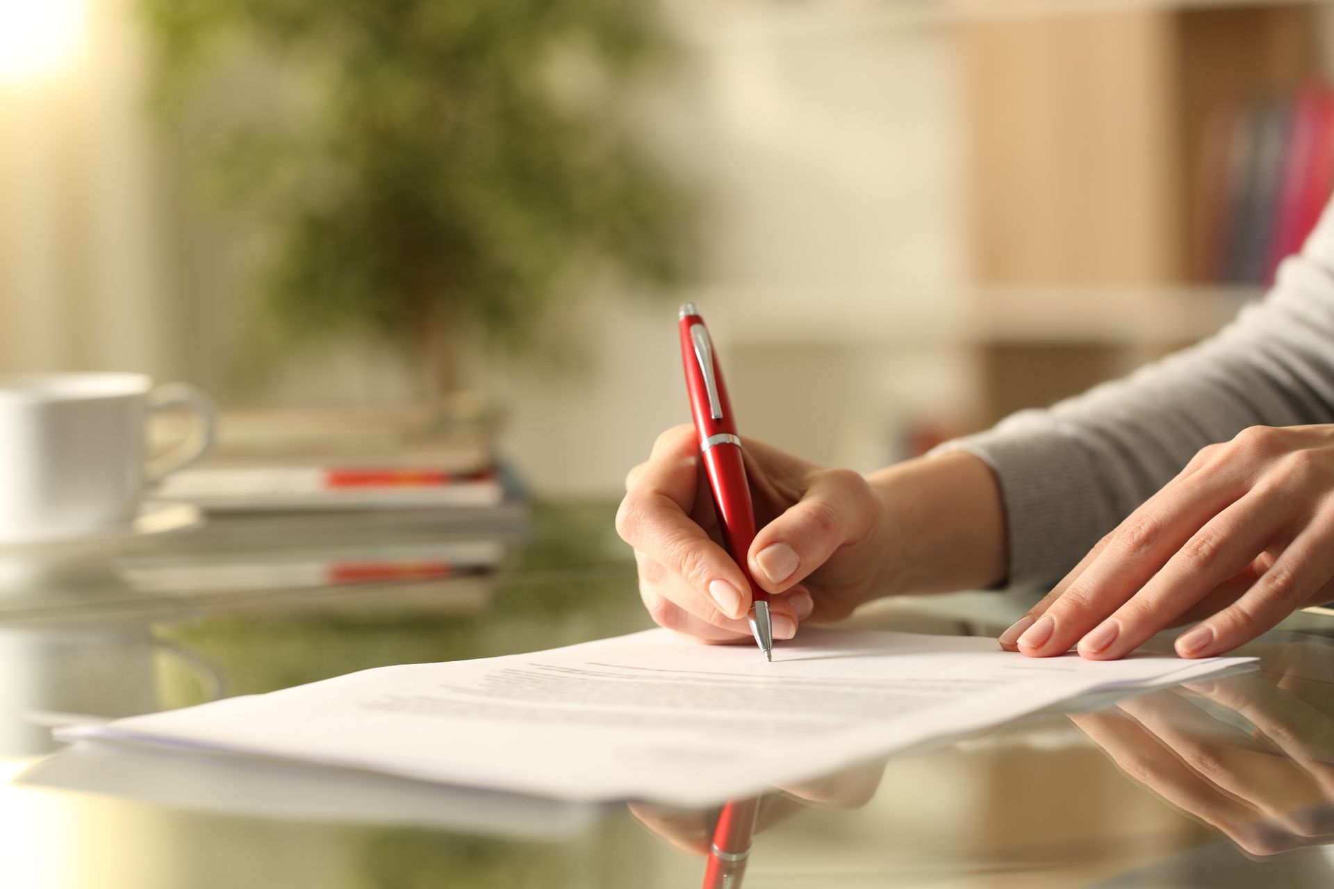 Person's hand holding a red pen, writing on a white document on a glass table.