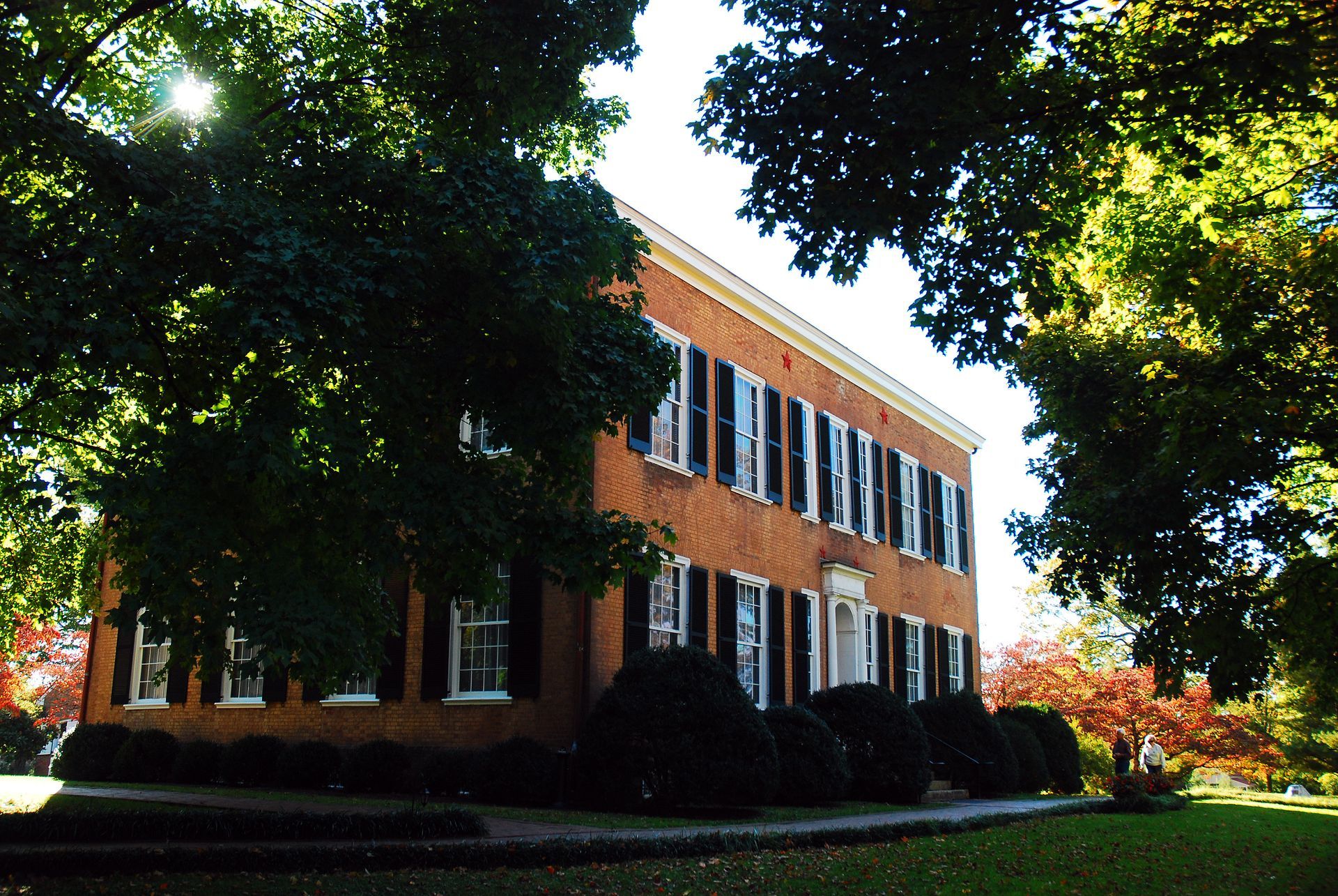 Two-story brick building with white trim, black shutters, and trees in a sunny setting.