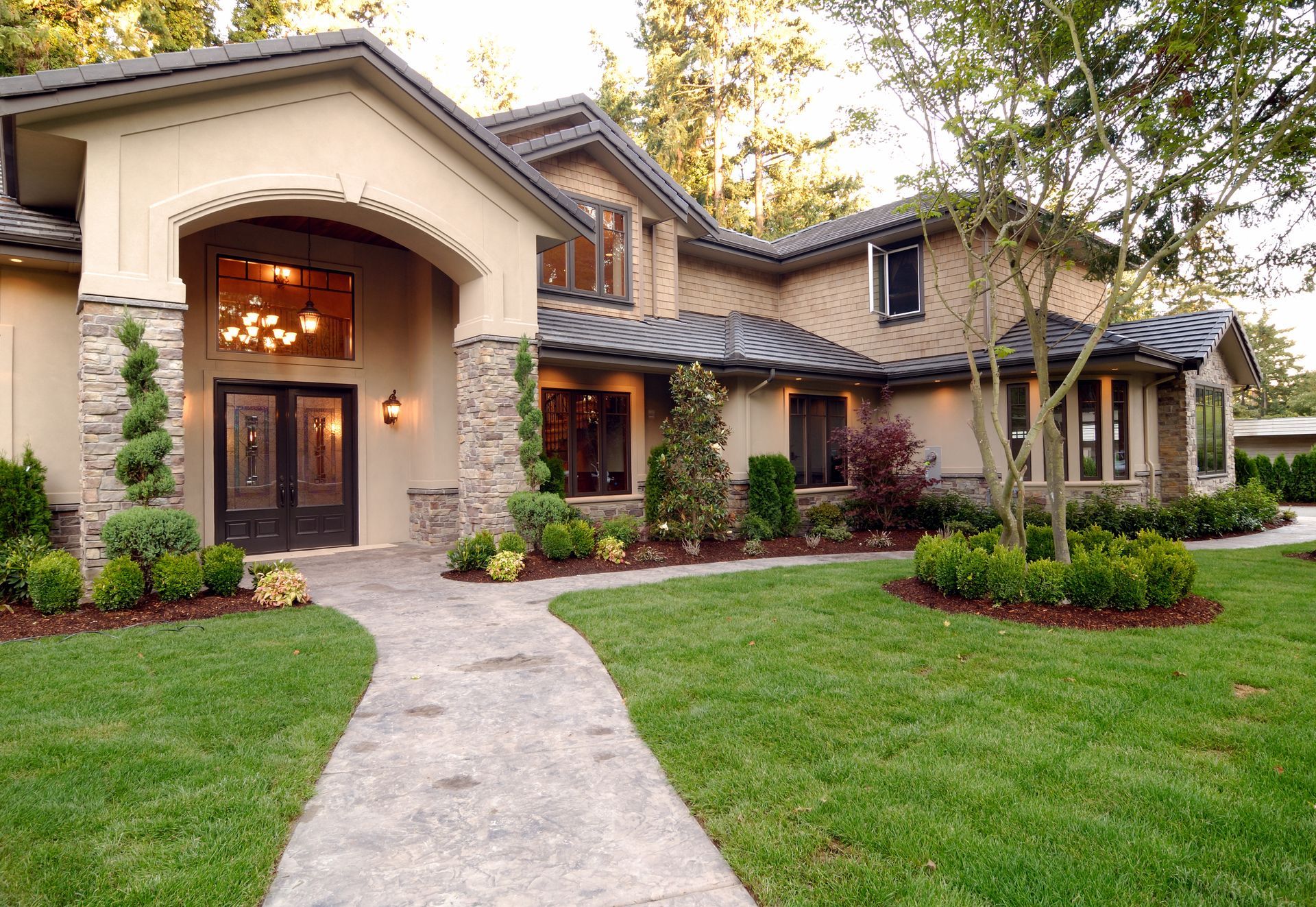 Beige and stone-faced house with arched entryway and manicured lawn. Concrete path leads to double doors.