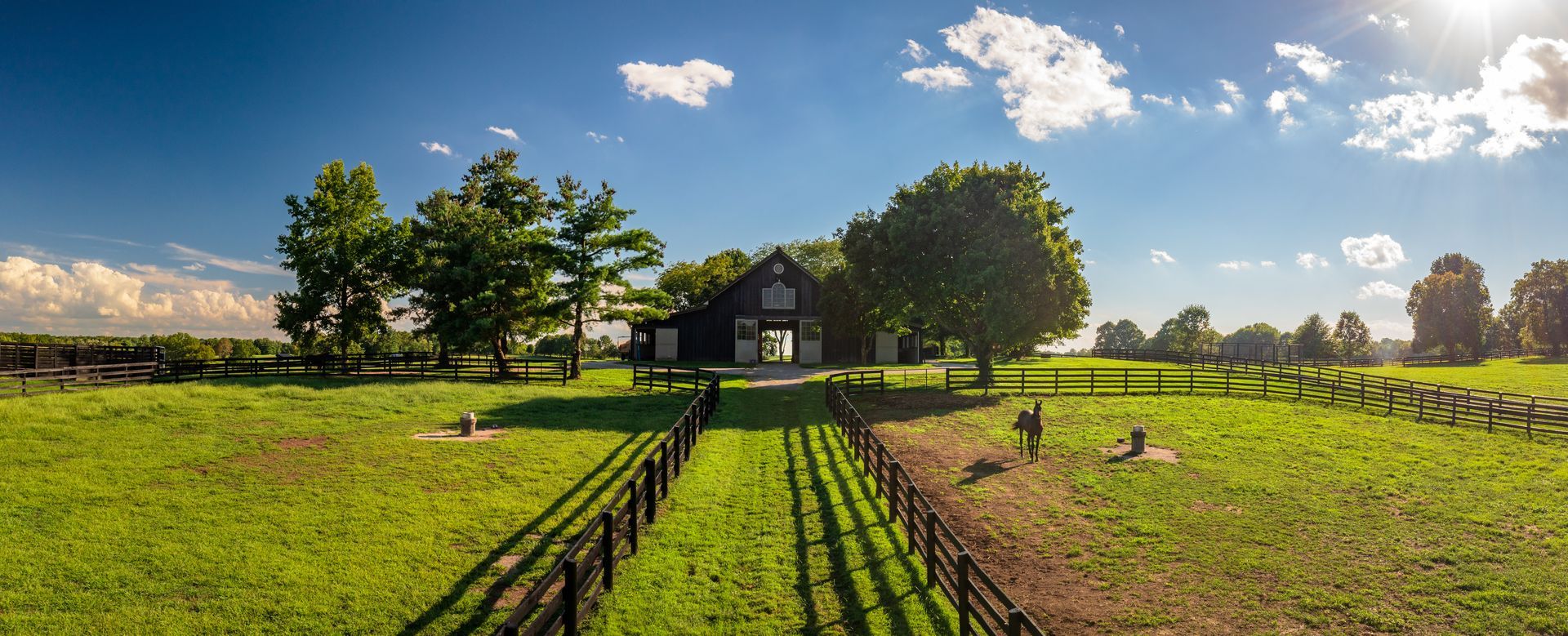 Green field with a barn, trees, and fence under a blue sky with clouds.