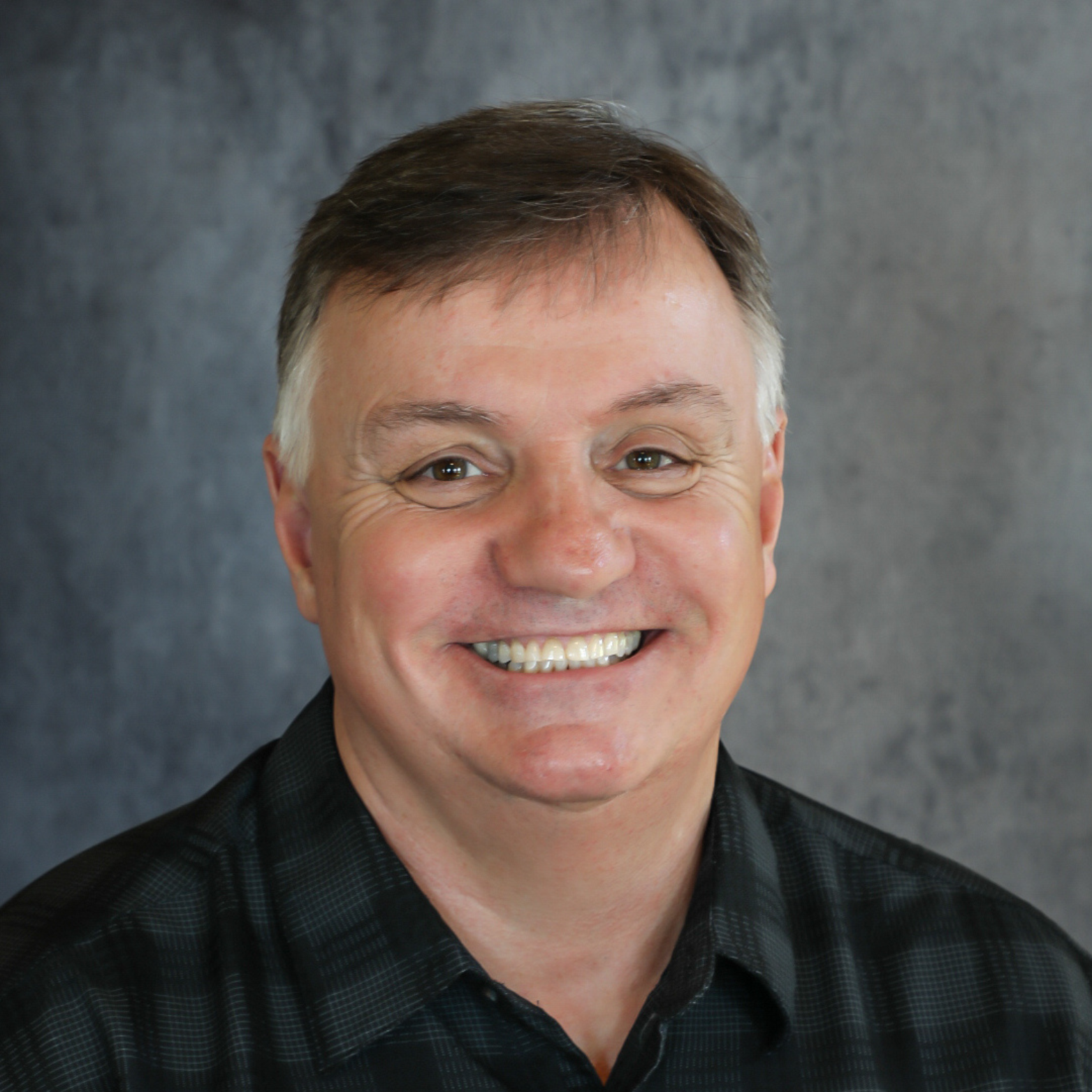Smiling man with short dark hair, wearing a black patterned shirt, in front of a gray backdrop.