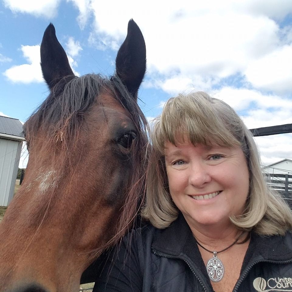 Kathleen Walls smiling next to a brown horse outdoors on a sunny day.