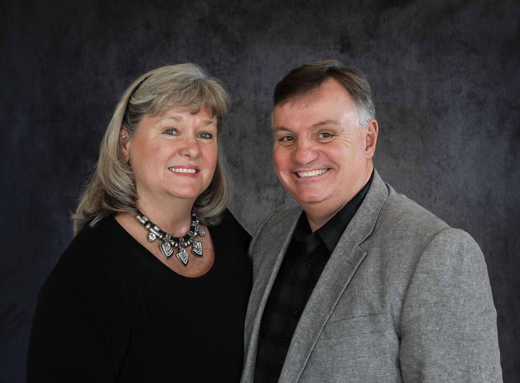 Smiling man and woman pose together; woman wears black top and necklace, man in gray blazer.