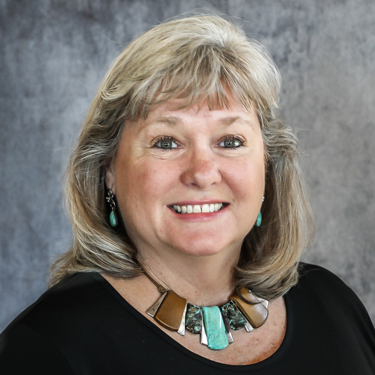 Woman with blonde hair wearing a necklace, earrings, and black top smiles at the camera.