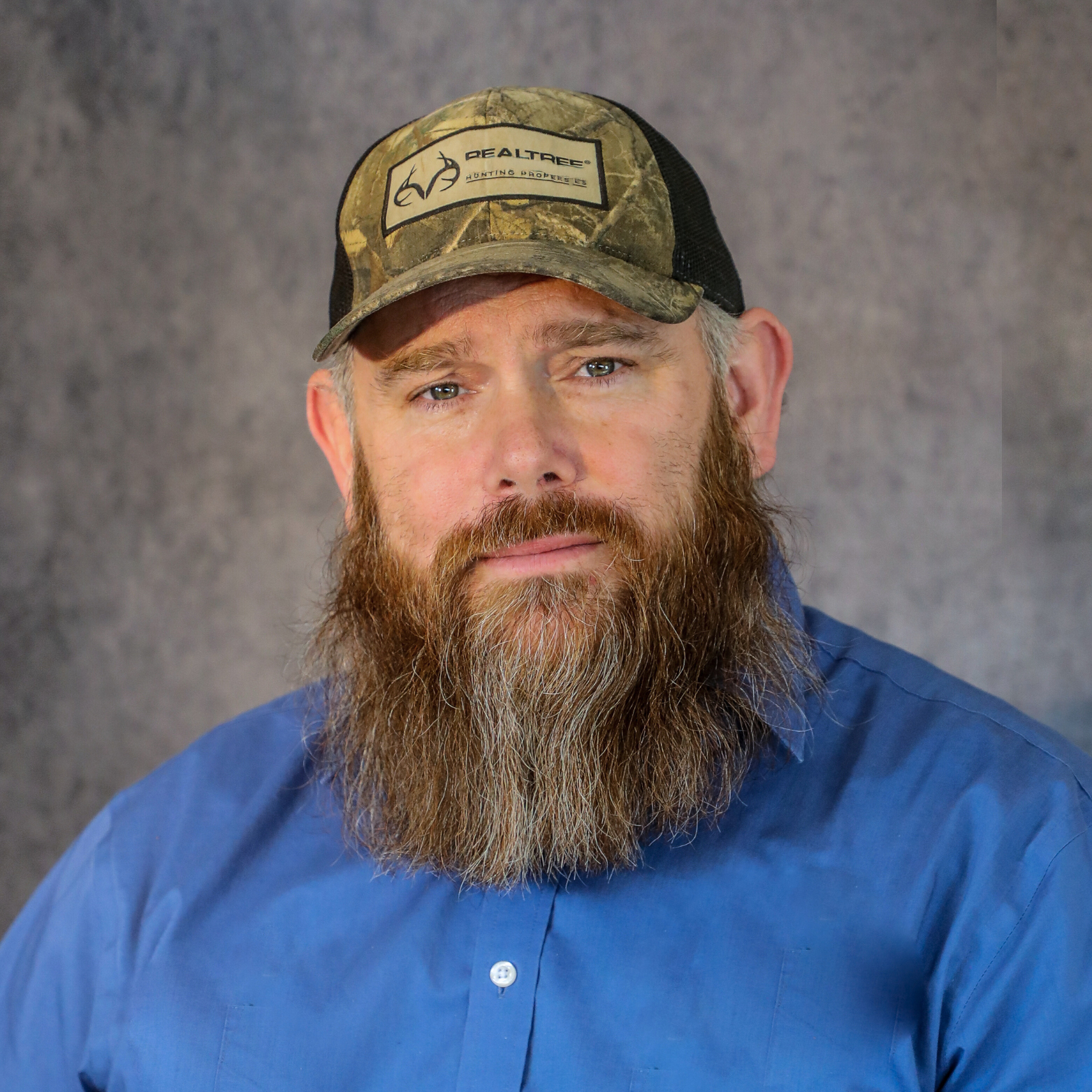 Man with a full beard and camouflage baseball cap wearing a blue shirt.