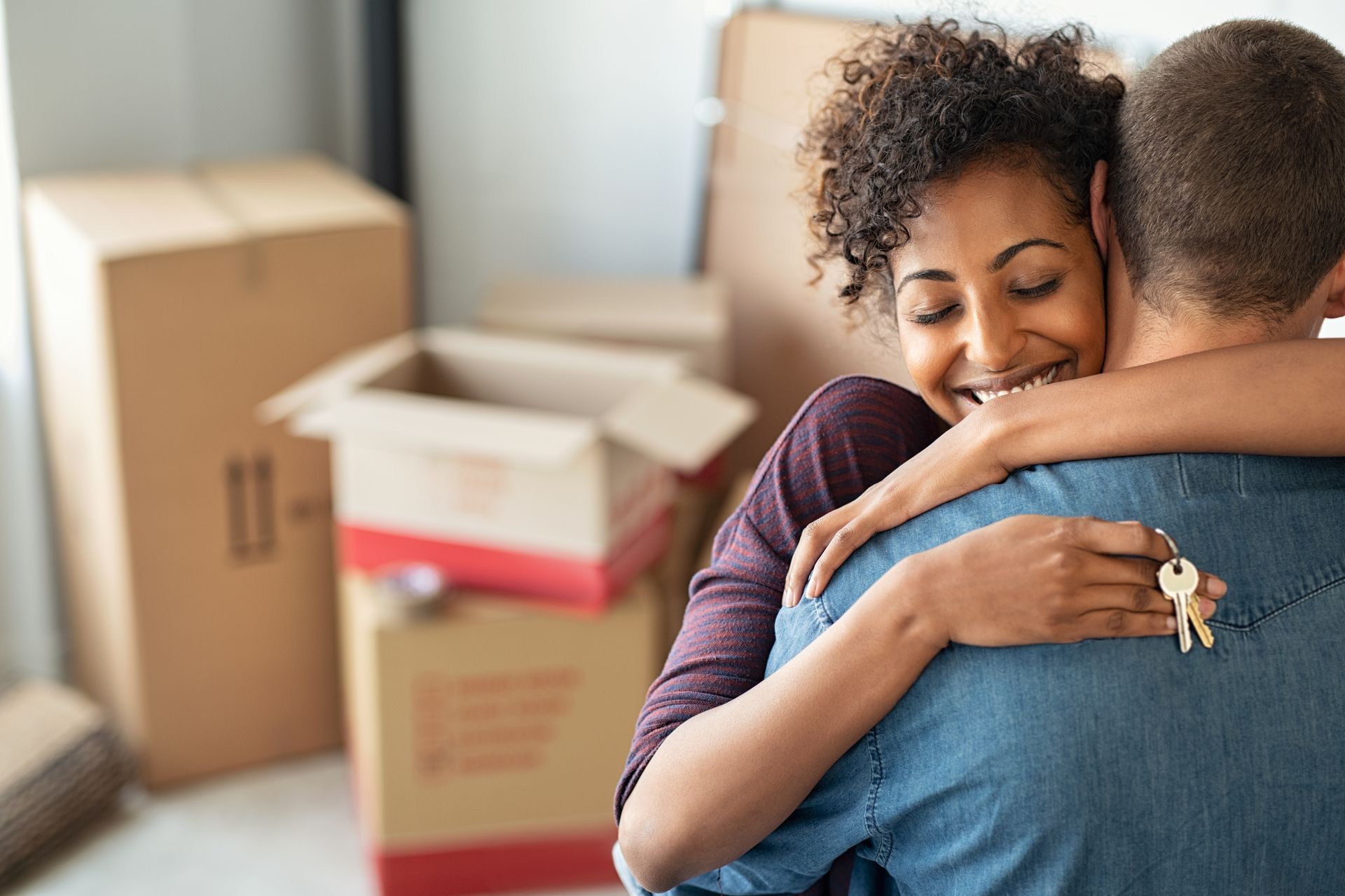 A couple embracing while holding keys to their new home with moving boxes in the background