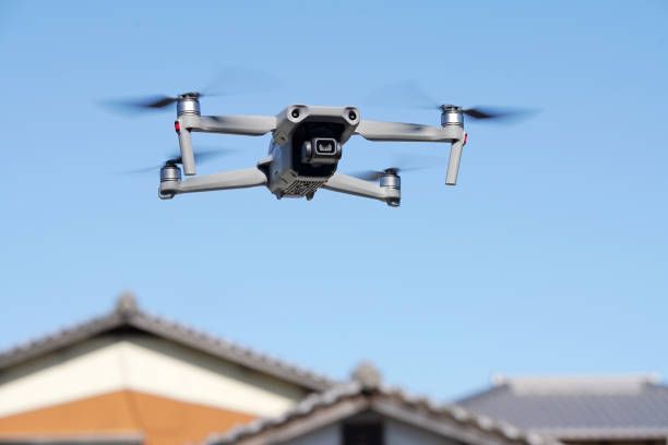 Drone flying in a blue sky above rooftops; gray body with spinning propellers and a visible camera.