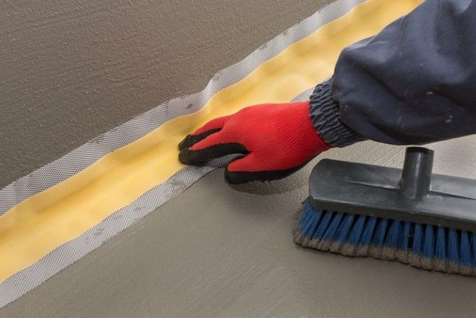 Gloved hand smoothing gray floor sealant beside yellow masking tape and a scrub brush
