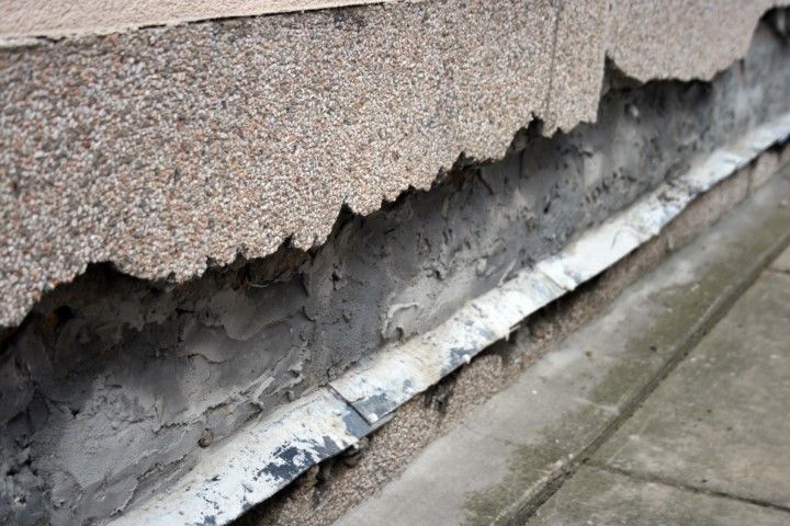 Cracked exterior wall with peeling stucco and exposed damaged layers along a sidewalk