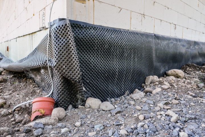 Black drainage fabric covering a house foundation on a rocky slope, with a red bucket nearby