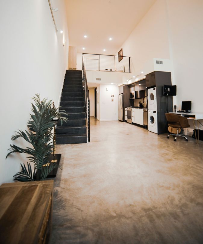 Modern loft-style room with high ceiling, staircase, plants, and desks along the right wall