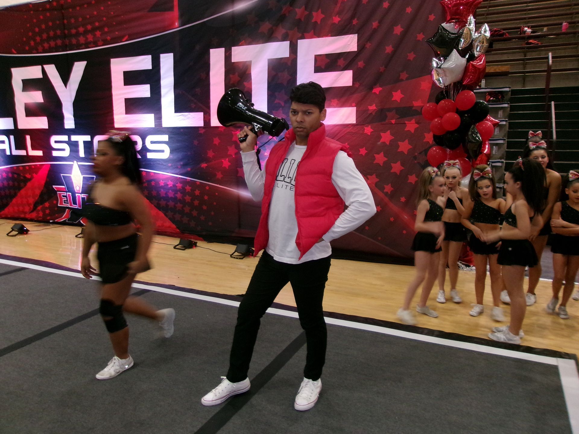 A person in a red vest holds a megaphone on a gym floor, with dancers and cheerleading props in the background.