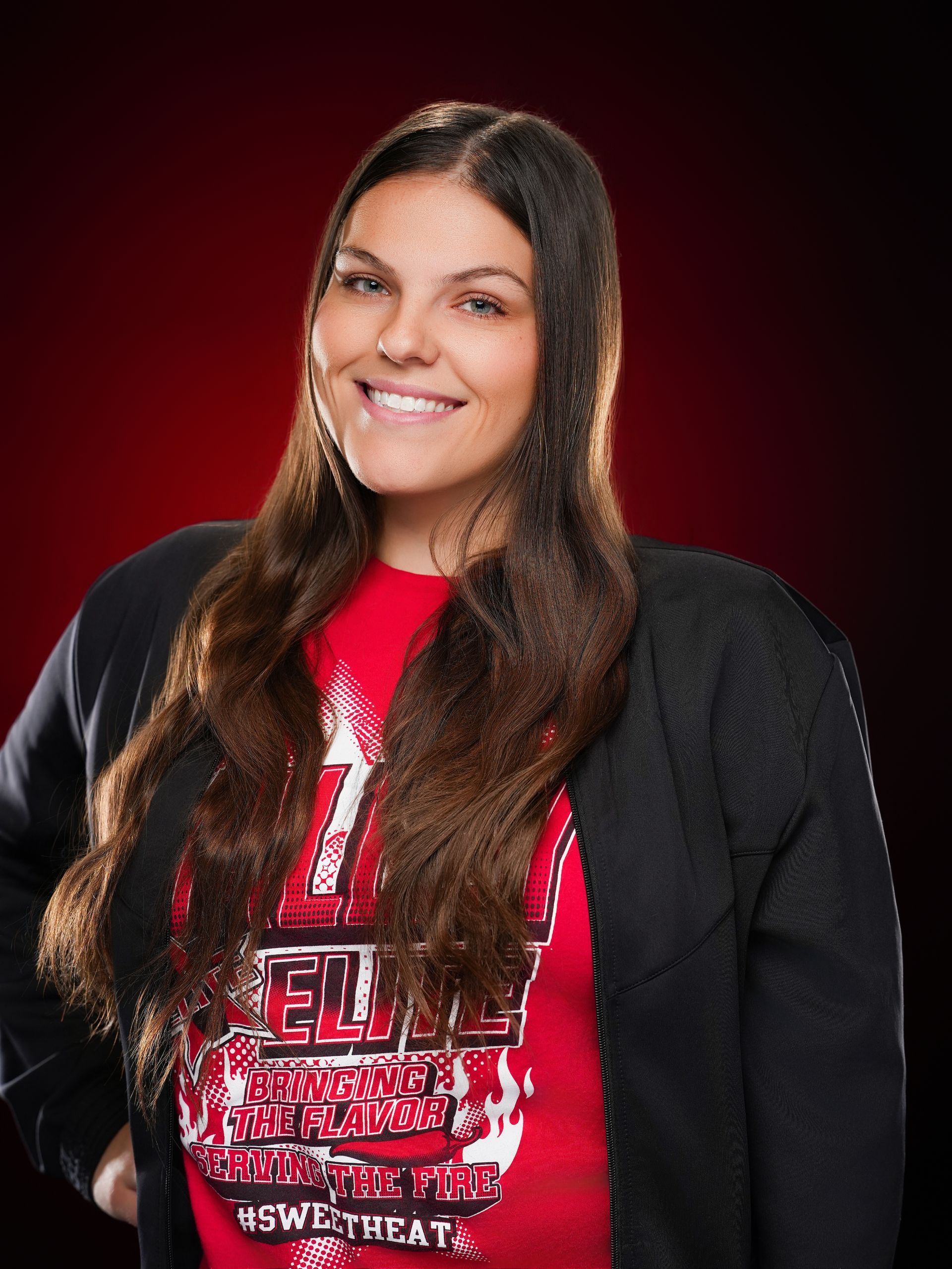 A smiling person with long dark hair wears a black jacket over a red graphic t-shirt against a dark red background.