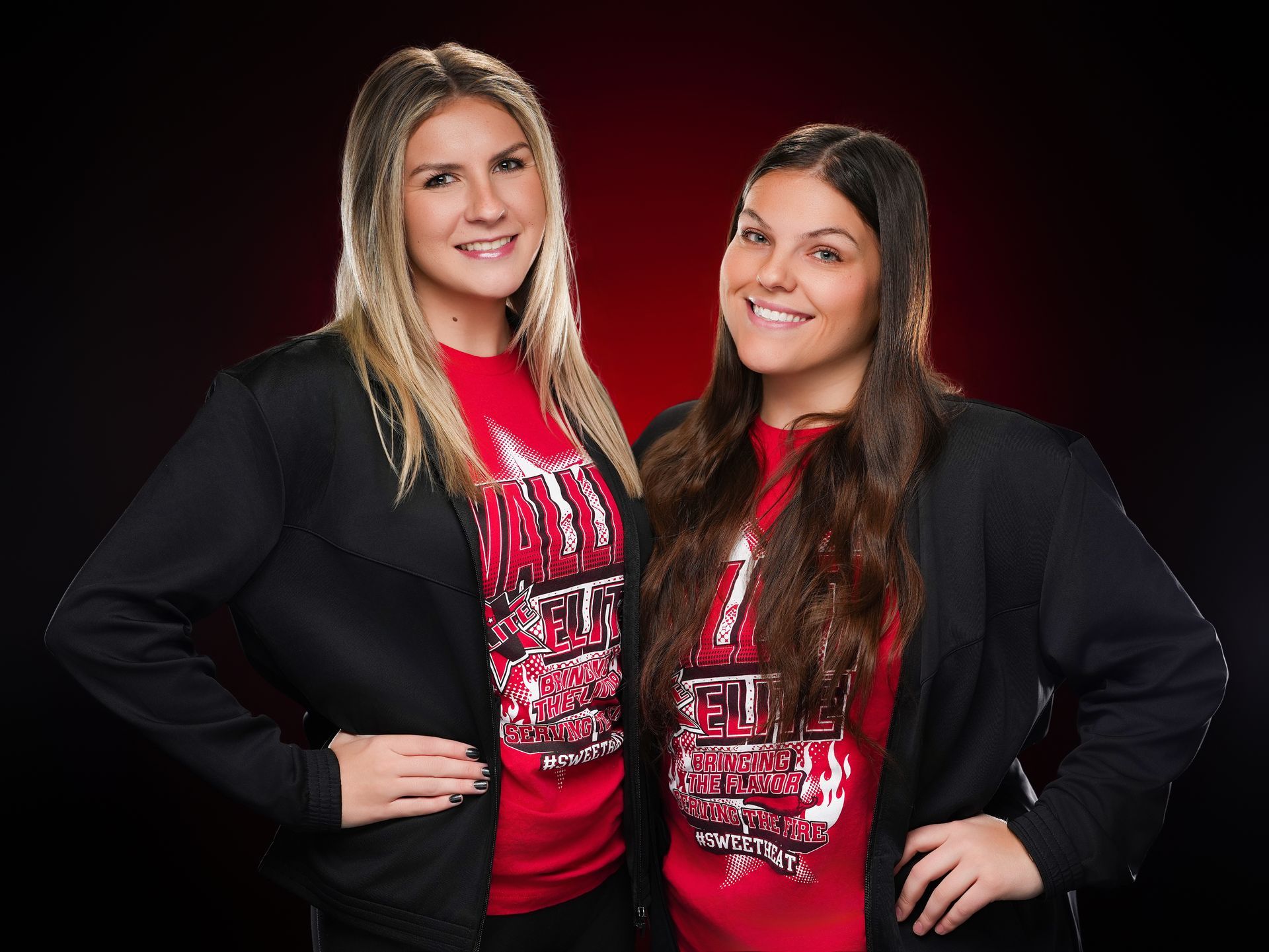 Two people wearing black jackets over red graphic t-shirts stand smiling against a black and red background.