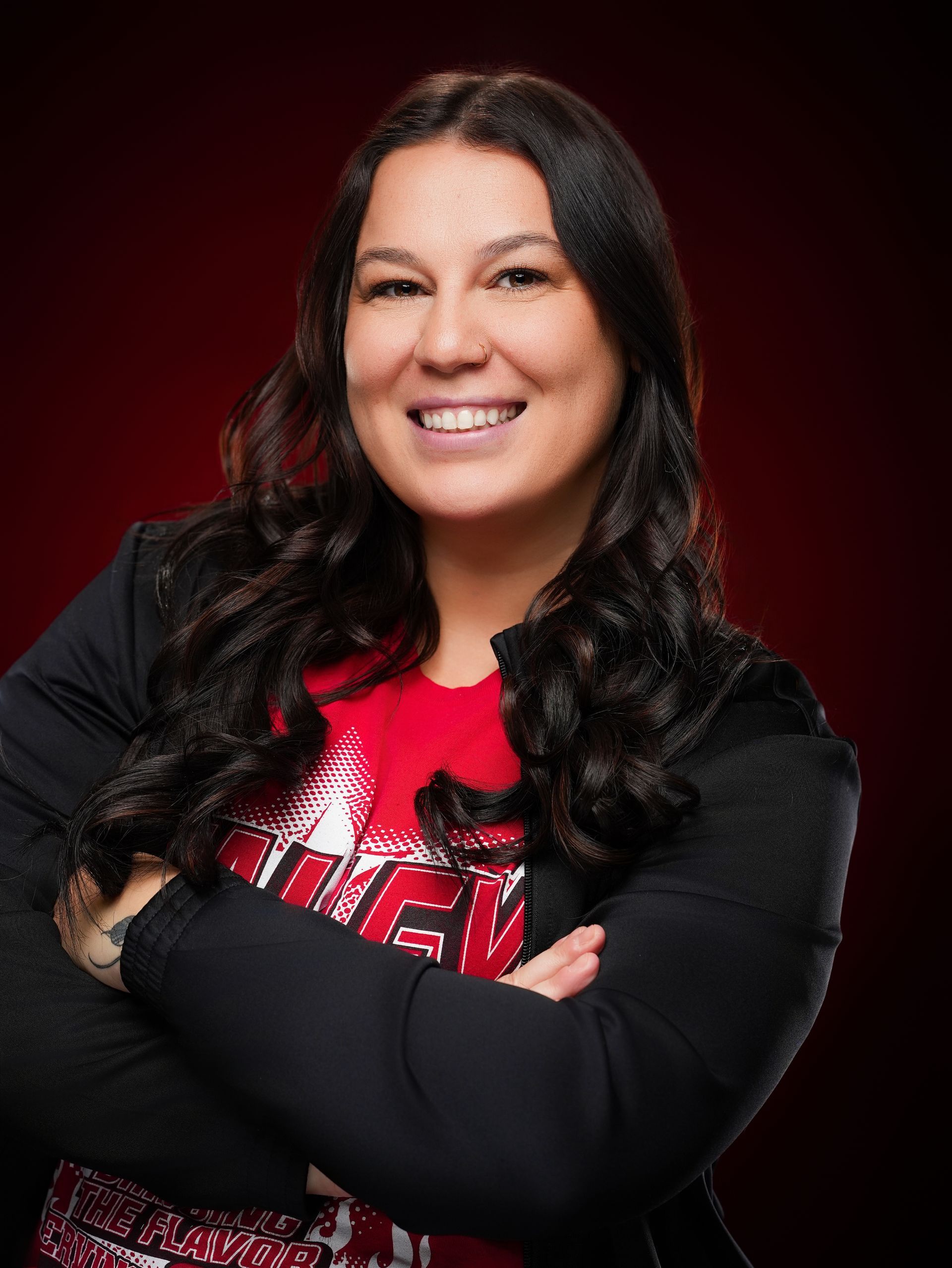 Smiling person with long, dark, wavy hair and arms crossed, wearing a red top and black jacket against a dark red backdrop.