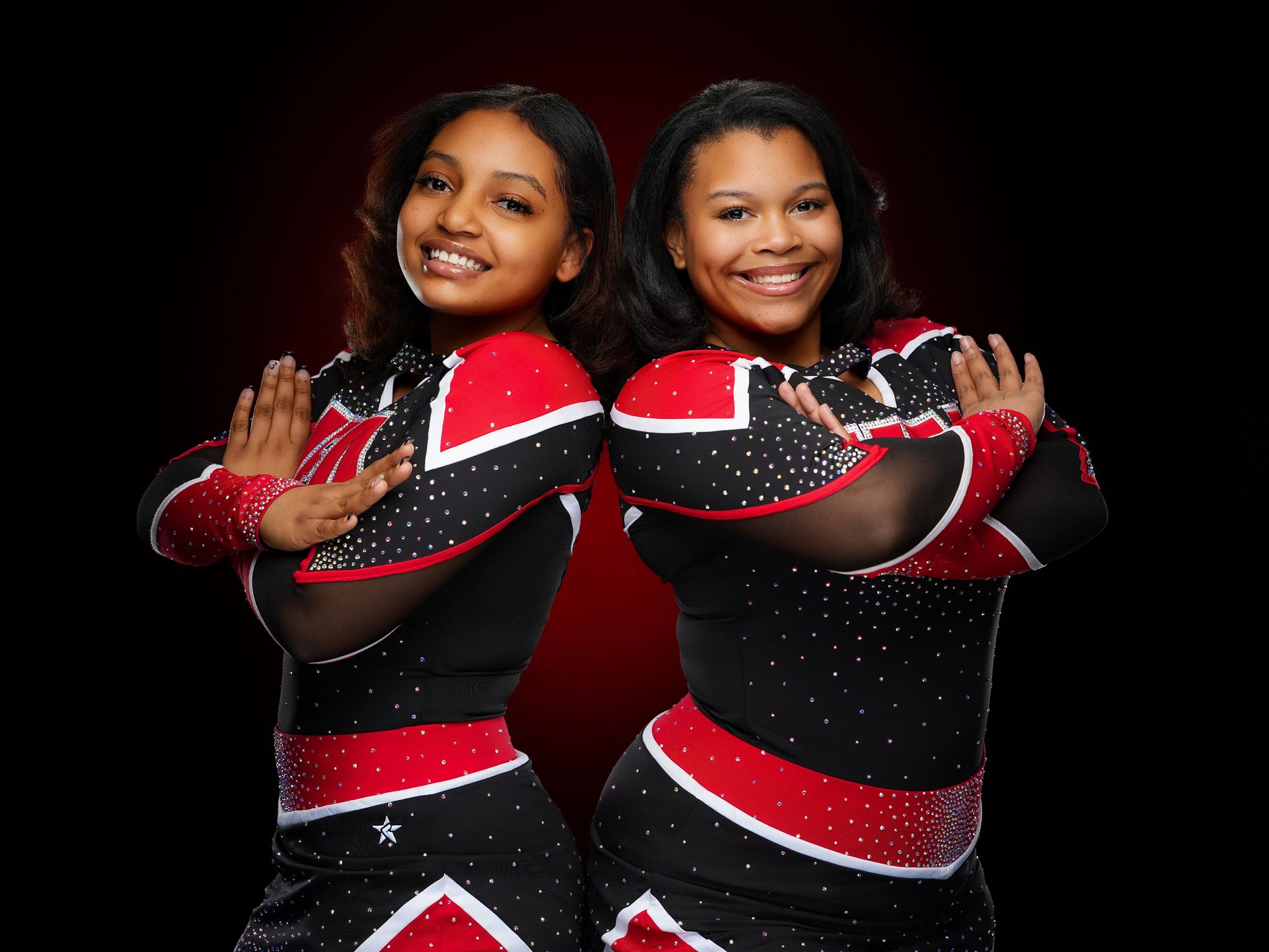Two smiling cheerleaders in black and red uniforms stand back-to-back with arms crossed against a dark background.