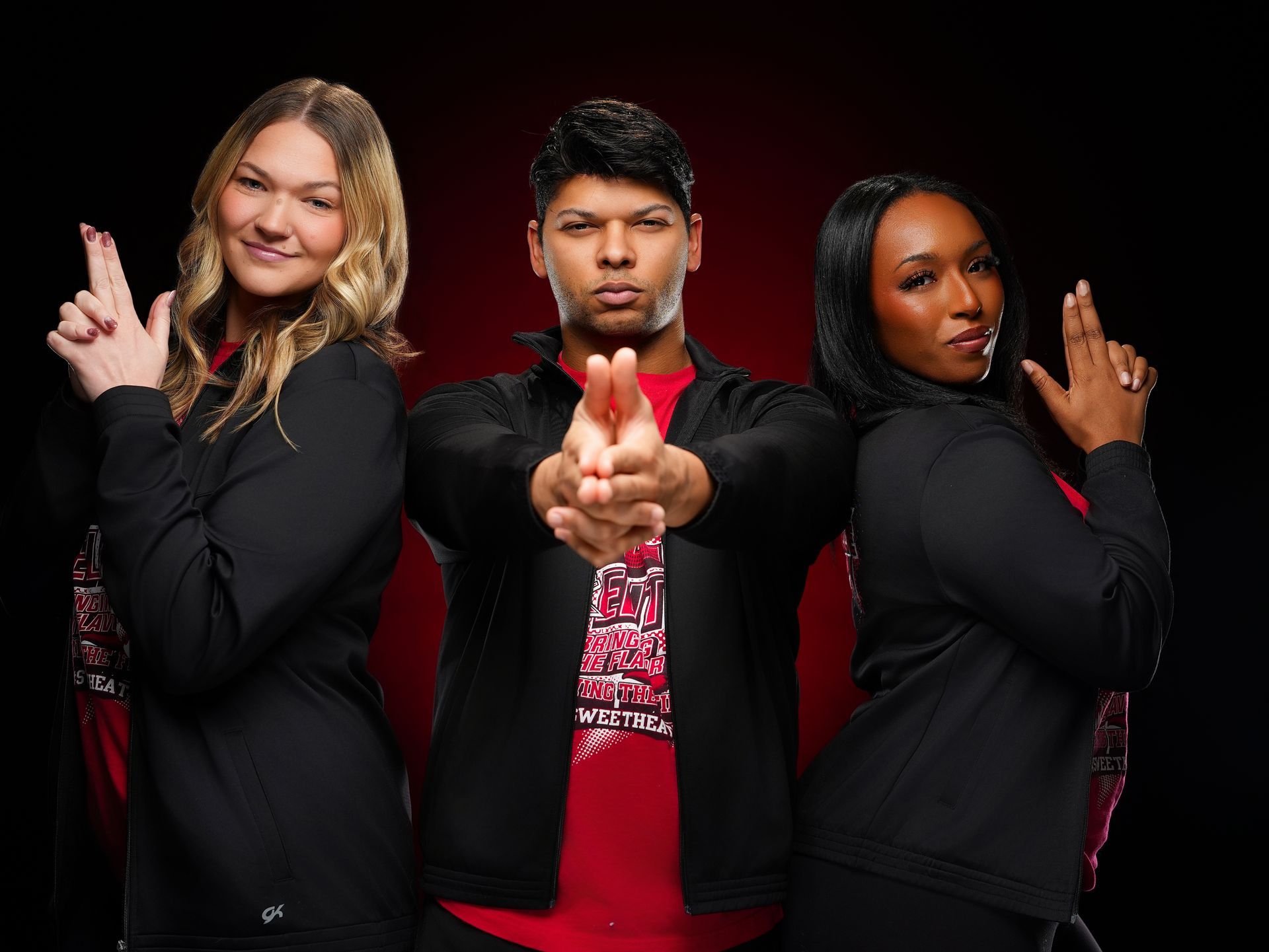 Three people wearing black zip-up jackets over red shirts stand against a dark red background, making finger-gun gestures.